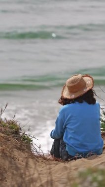 Diz çöküp kumulların arasında kumların üzerinde dizleri üzerine çökmüş, hasır şapka takmış, arka plandaki bulanık denize bakan bir kadının dikey videosu. Refah, sakinlik, içgözlem ve yavaş seyahat kavramı