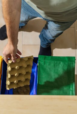 Evcil bir mutfaktaki mavi bir geri dönüşüm kutusuna karton yumurta kutusunu koyan bir adamı gösteren mesajlar için yer ile dikey genel bakış. Atık ayırma ve sürdürülebilirlik.