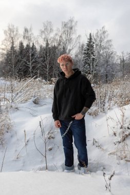 Pembe sarı saçlı, gözlüklü ve siyah kapüşonlu genç modellerin portresi. Kışın karlı havada, ormanın yanındaki tarlada duruyordu.