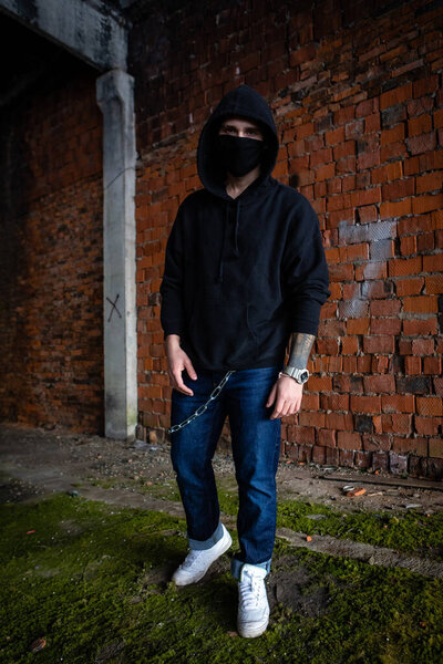 Молодой привлекательный гангстер-модель в черной маске против ковидного коронавируса. Протест в капюшоне или толстовке возле оранжевой кирпичной стены. Сила, агрессия и готовность к бою. городская жизнь