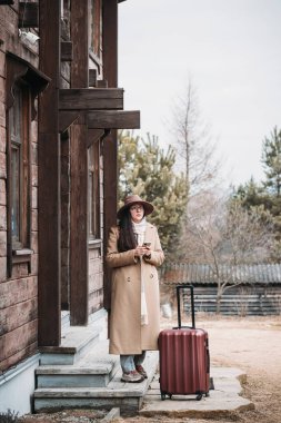 Otuzlarında bir kadın, kırsal kesimde kırsal ahşap otelin yanında valiziyle seyahat ve eğlence konseptiyle geziyor. Kadın taksi bekliyor.
