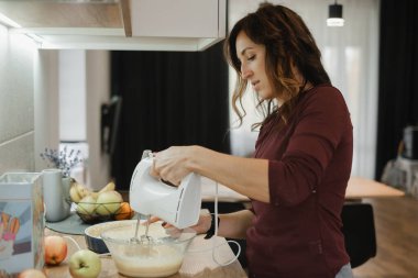 Genç bir bayan taze meyve ve sebzelerle, sağlıklı yemek hazırlama ve karıştırma malzemeleriyle parlak mutfakta turta hazırlıyor.
