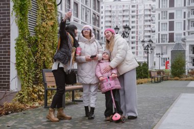Mutlu kadınlar ve küçük kızlar, modern konut binalarının yanında, rahat kabanlar ve şapkalar içinde açık hava selfie 'si için birlikte poz veriyorlar.