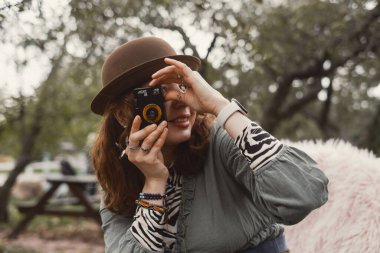 30 'lu yaşlarındaki kadın, açık hava parkında şapkası ve katmanlı boho kıyafeti giymiş, küçük bir film kamerasıyla çekim yaparken gülümsüyor. Yaratıcı hobi