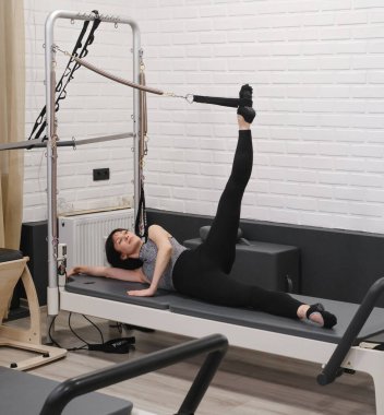 40 'lı yaşlarda bir kadın pilates kulesi egzersizi yapıyor. Kablo direnciyle, yenilikçi paspasın üzerinde yatarak bacak kaldırma egzersizi için kayışlar kullanıyor.