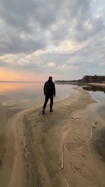 Bulutlu bir bahar gününde kumlu bir plajda tek başına yürüyen bir adam sakin suya yansıyor. Dikkatli bir yürüyüş, düşünme ve dinlenme.