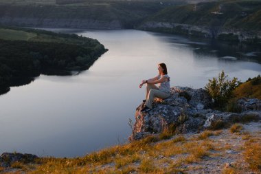 Young woman traveler sits on rocky cliff edge enjoying panoramic view of winding river canyon during golden hour sunset in summer nature.