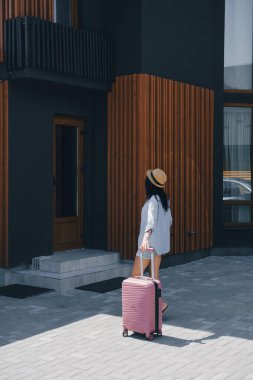 Şapkalı genç bayan gezgin modern apartman girişine doğru pembe bavulunu çekiyor. Yaz tatili ve konaklama konsepti.