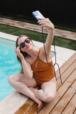 Turuncu mayo ve güneş gözlüklü mutlu genç kadın yaz tatilinde ahşap güvertede mavi havuz kenarında oturup akıllı telefonuyla selfie çekiyor..