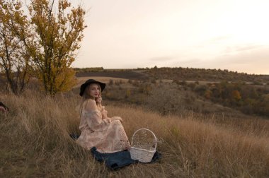 Akşamları tepede çimlerin üzerinde oturan şapkalı ve elbiseli çekici genç bir kadın.