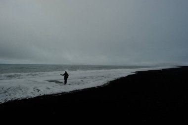 Kumsalda sırt çantası ve şapkası olan siyah ceketli bir kadın.