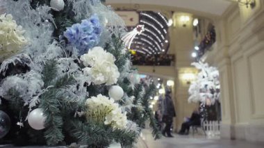 Dekoratif çiçeklerle süslenmiş Noel ağacı, geçen ziyaretçilerin arka planında, alışveriş merkezinde duruyor. Sabit kamera