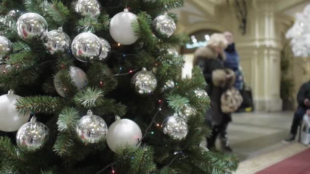 Επισκέπτες περνούν πίσω από ένα χριστουγεννιάτικο δέντρο διακοσμημένο με ασημένιες μπάλες σε GUM — Αρχείο Βίντεο