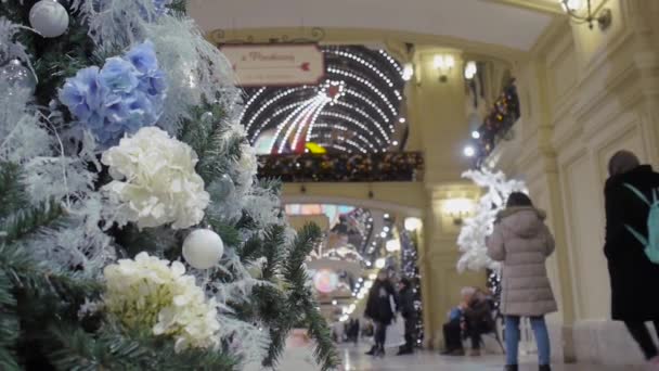 Dekoratif çiçeklerle süslenmiş Noel ağacı, geçen ziyaretçilerin arka planında, alışveriş merkezinde duruyor. — Stok video