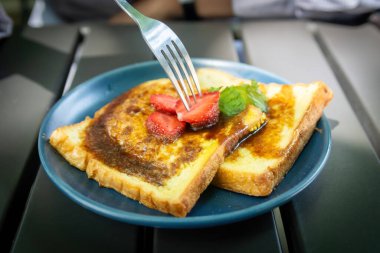 Taze çilek kesen bir çatal. Altın Fransız tostu dilimleri üzerine. Şerbetle serpiştirilmiş, koyu tabakta.