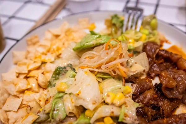 Zengin dilimlenmiş sığır eti, taze karışık salata, mısır ve beyaz tabakta gevrek tortilla cipsi içeren dengeli bir füzyon tabağı..