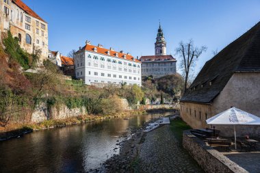 Cesky Krumlov, Çek Cumhuriyeti - 23 Kasım 2025: Vltava Nehri üzerindeki antik bir kasaba. Yeşil ağaçlar arasında nehir kıyısında konforlu renkli evleri olan resimsel bir manzara. Güneşli gün mavi gökyüzü bulutları.