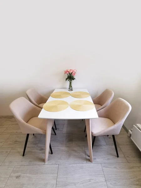 Small kitchen corner with white walls, a dining table and beige armchairs. Vase with roses as an element of interior design on the table in the room. Interior of a modern apartment in white and beige