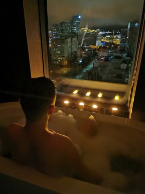 Genç adam mum ışığında pencerenin yanındaki köpük banyosunda banyo yapar ve dinlenir. Dinlenme ve rahatlama, spa tedavileri.