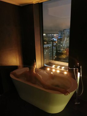 Genç adam mum ışığında pencerenin yanındaki köpük banyosunda banyo yapar ve dinlenir. Dinlenme ve rahatlama, spa tedavileri.