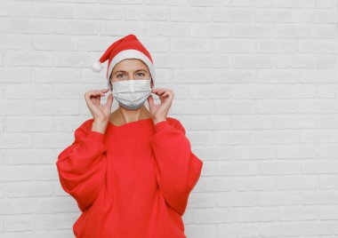 Noel Baba şapkalı güzel bir kadın model tıbbi maske takıyor. Noel ve yeni yıl kutlamaları salgın sırasında. Dikkatli davran..
