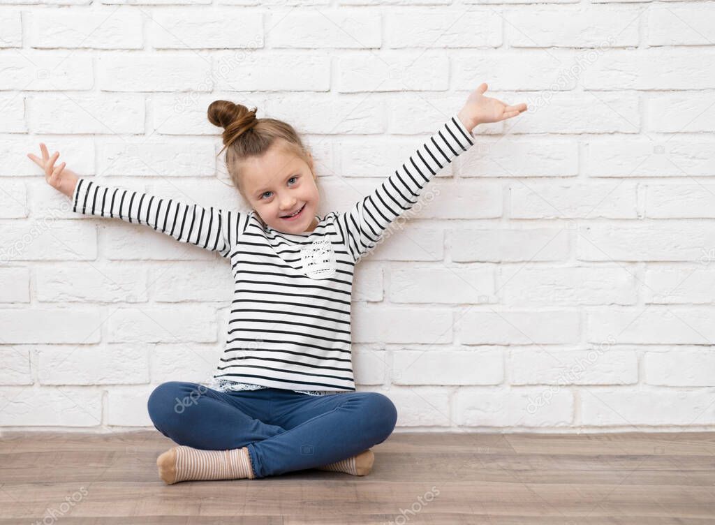 A happy little Caucasian girl of 5-6 years old is sitting on the floor against a white brick wall with her arms outstretched. The concept of a happy childhood, place for text