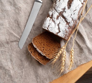Tahta bir tahtanın üzerinde buğday kulakları ve keten peçetenin üzerinde bir bıçakla kesilmiş ev yapımı koyu kokulu taze ekmek. Kırsal ev yapımı konsepti. Yukarıdan görünüşü