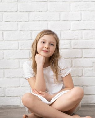 Mutlu bir çocuğun portresi. 8-9-10 yaşlarında küçük sarışın bir kız. Yerde, boş beyaz bir duvarın arkasında oturuyor. Ortam planı