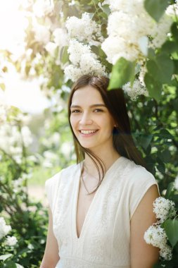 Portrait of a smiling beautiful young Caucasian brunette girl against a background of white blooming lilac on a warm sunny day. Flowers in your hair. Copy space.
