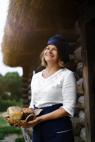 a woman baker with a basket of bread in her hands in a blue baker\'s cap and apron against the background of a wooden log bakery. Yeast-free wheat and rye bread with sourdough. Eco bakery.