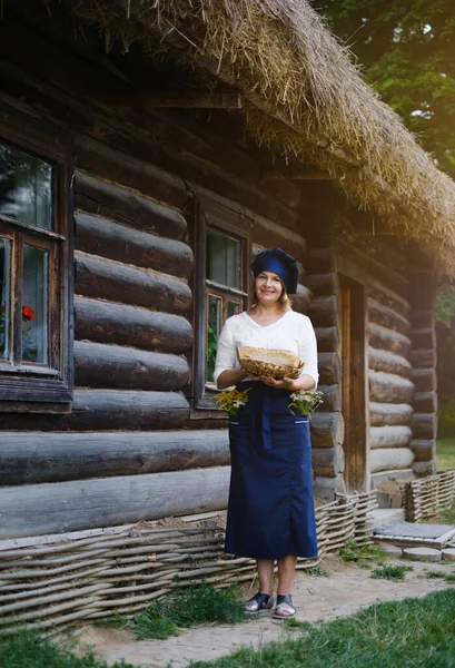 a full-length woman baker with a basket of bread in her hands in a chef\'s hat and apron against the background of a wooden log village house. Yeast-free wheat and rye bread with sourdough. Eco bakery.