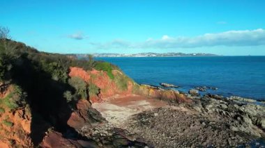 Broadbay, Torbay, Devon, İngiltere: DRONE VİEWS: Uzakta Torbay denizi ve Torquay kasabasını ortaya çıkarmak için kızıl kumtaşı uçurumlarının üzerinden yükselmeden önce deniz kıyısı kayaları üzerinde alçaktan uçar. Güney Devon, İngiltere 'nin popüler tatil beldesidir..