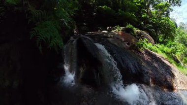 Yeşil orman bitkileri ve güneş ışığıyla çevrili karanlık kayaların üzerinde akan şelale. Dauin, Zenci Doğulu. Filipinler.