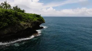 Bulutlu bir gökyüzünün altında mavi denizin üzerinde yeşil bitki örtüsüyle kaplı uçurum manzarası. Siargao, Filipinler.