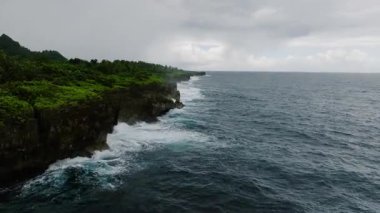 Dik kayalıklar, bulutlu gökyüzünün altında, üzerinde orman olan okyanus dalgalarıyla karşılaşıyor. Siargao, Filipinler.