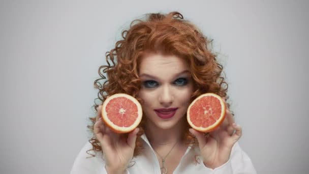 Женщина с половинками грейпфрута. Веселая девушка кусает губы на сером фоне — стоковое видео