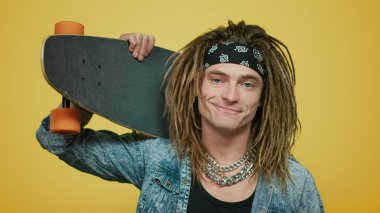 Omzunda kaykay tutan bir adam. Sarı arkaplanda poz veren kaykaycı