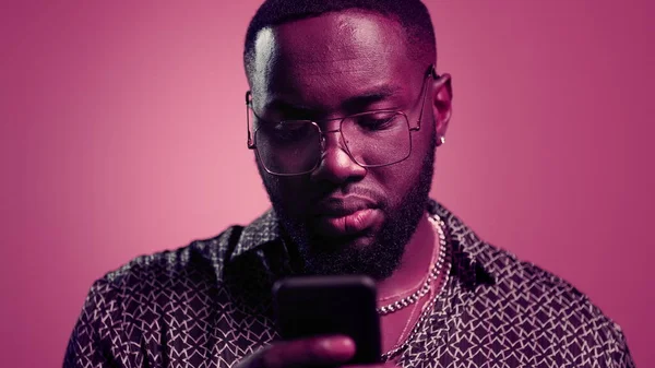 Afro czyta wiadomości w studio. Mężczyzna za pomocą smartfona na różowym tle — Zdjęcie stockowe
