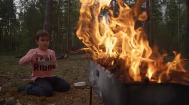 Barbekü ızgarasından çıkan parlak alevler ateşin yanında oturan çocuğun siluetini aydınlatıyor.