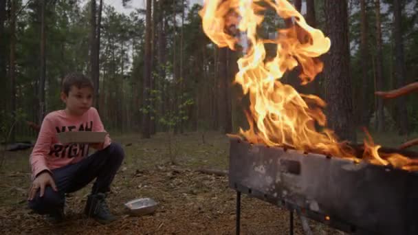 Handsome teenage boy eating fast food near barbecue bonfire in forest. — Stock Video