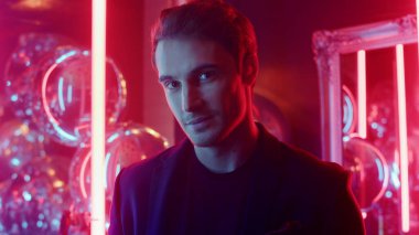 Gece kulübünde kameraya bakan utangaç adam. Neon arka planda poz veren erkek kişi