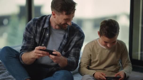 Πατέρας και γιος gaming κινητά τηλέφωνα. Ο μπαμπάς και το αγόρι κατασκοπεύουν ο ένας τον άλλον. — Αρχείο Βίντεο