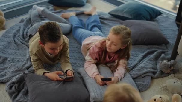 Kinderen die smartphones spelen. broers en zussen liggend met mobieltjes volgende kind op tapijt. — Stockvideo