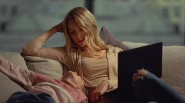 Bilgisayarlı anne evde kızıyla sohbet ediyor. Anne kız yüzlü kanepeye dokunuyor