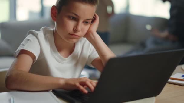 Smutny dzieciak uczy się zdalnie w domu. Zmęczony chłopiec siedzi przy stole z komputerem.. — Wideo stockowe