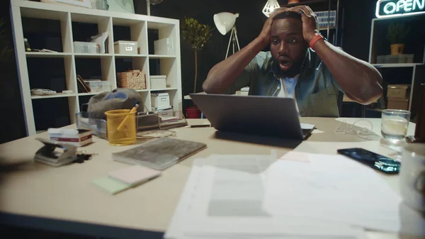 Frustrated african businessman grabbing head in dark coworking.