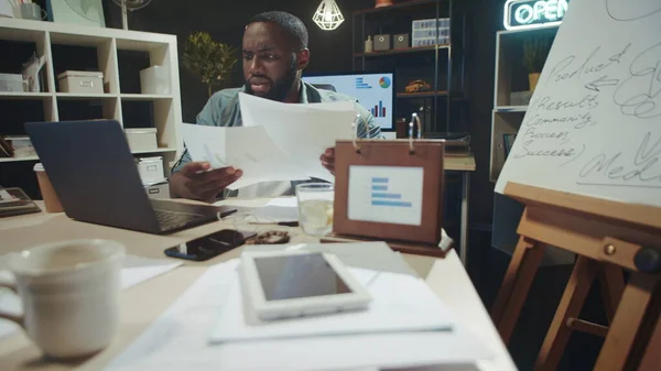 Stressed afro guy checking data on laptop screen with documents indoors.