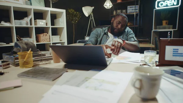 Porträt eines Afroamerikaners, der im dunklen Büro mit Papieren in Laptopnähe arbeitet. — Stockfoto