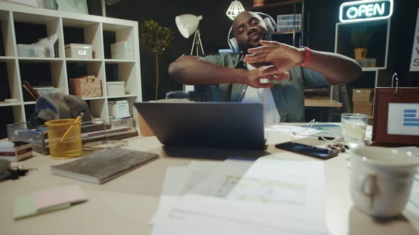 Portrait of afro man having fun in headphones near laptop in dark office.