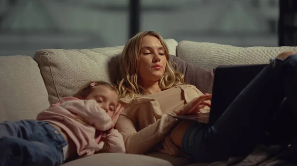 Mom using laptop next to daughter on couch. Girl falling asleep mom shoulder